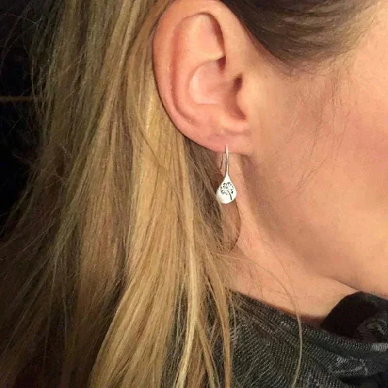 Silver teardrop earrings worn on ear, engraved dandelion line art on curved face